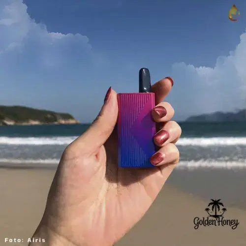 Mano sosteniendo la batería Airis Mistica 3 en la playa, con mar y cielo en el fondo, destacando su diseño colorido.